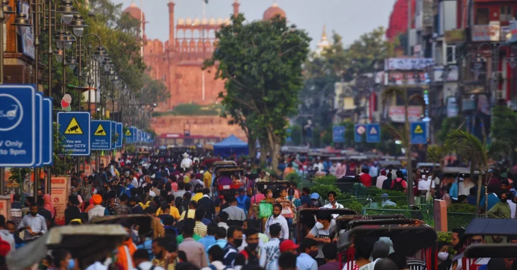 Crowded street view of Chandni Chowk Delhi with people, rickshaws, shops, and Red Fort in the background.